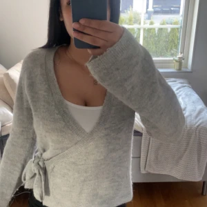 Ljusgrå omlottkofta med knyt - Mysig ljusgrå omlottkofta med knyt i sidan och v-ringning. Tröjan har långa ärmar och är perfekt för lager-på-lager. Mjuk och skön känsla, passar till många olika outfits.