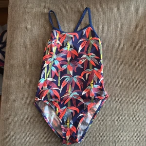 Färgglad baddräkt från Funkita - Säljer en baddräkt från Funkita med tropiskt blommigt mönster i rött, grönt, blått och gult på mörkblå botten. Modellen har smala axelband och öppen rygg. Perfekt för simträning eller strandhäng.