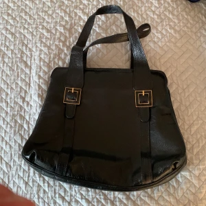 Svart handväska med gulddetaljer - Stilren svart handväska i glansigt material med två axelband och snygga fyrkantiga guldfärgade spännen framtill. Perfekt storlek för vardagens essentials och ger en klassisk look. Osäker vilket märke det är men det är i ett jättefint skinn och kvalitet 