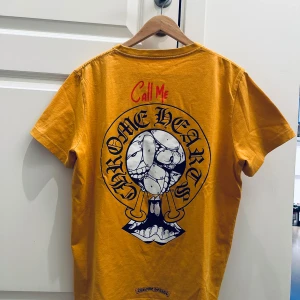 Call me logo Gul t-shirt från Chrome Hearts - Gul t-shirt från Chrome Hearts med tryck på ryggen och bröstet. Stor logga och texten 'Call Me' i rött på baksidan, samt liten bröstficka framtill. Rund halsringning och korta ärmar. Perfekt för dig som gillar streetwear och statement-plagg.