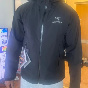 Svart skaljacka från Arc'teryx - Säljer en svart skaljacka från Arc'teryx med huva och vattentät dragkedja. Jackan är tillverkad i Gore-Tex-material och har två sidofickor med dragkedja. Perfekt för utomhusaktiviteter och skyddar mot regn och vind.