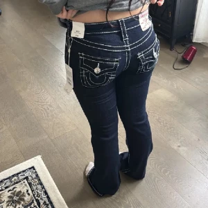 Mörkblå jeansbyxor från True Religion - Snygga mörkblå jeans från True Religion med vita kontrastsömmar och dekorativa fickor med knappar. Byxorna har en bootcut-stil och är lågmidjade, vilket ger en klassisk look. Perfekta för dig som vill ha en stilren och trendig outfit. Helt nya med lappar köpt för 1599kr❤️ midjemått ca 35 cm(stretchiga) innerbenslängd 80 cm