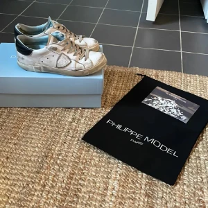 Vita sneakers från Philippe Model - Säljer ett par vita Philippe Model sneakers med grå detaljer och svarta hälpartier. Skorna har snörning och en klassisk låg profil med rund tå. Perfekta för dig som gillar stilrena och trendiga sneakers.