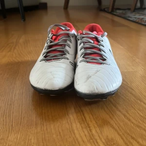 Nike fotbollsskor i vitt och svart - Säljer ett par Nike fotbollsskor i vitt och svart med röda detaljer. Har används i ungefär 2 månader men funkar exakt som nya. Skorna har lite smuts fläckar men fungerar ändå som ett par nya.