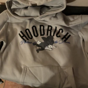 Grå hoodie från Hoodrich - Säljer en grå hoodie från Hoodrich med tryck både fram och bak. På framsidan står det 'Hoodrich' och på baksidan finns ett stort H med änglar och texten 'From Nothing To Something'. Hoodien har huva och magficka.