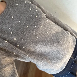 Grå stickad tröja med pärlor - Superfin grå stickad tröja med pärldetaljer framtill. Tröjan har rund halsringning och långa ärmar. Perfekt för att addera lite extra till din outfit. Mjuk och skön att bära.