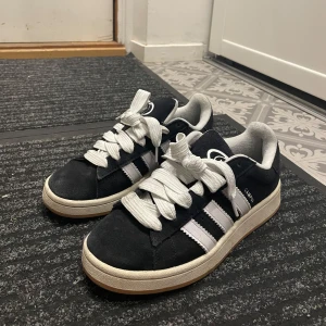 Svarta Adidas Campus sneakers med vita ränder - Säljer ett par klassiska Adidas Campus sneakers i svart mocka med vita ränder och vit sula. Skorna har rund tå, platt sula och snörning. Extra svarta skosnören medföljer. Perfekta för en stilren vardagslook. Använt ett par gånger.