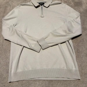 Grå pikétröja Next - Säljer en stilren beige pikétröja från Next med lång ärm och dragkedja vid halsen. Tröjan har klassisk krage och ribbade muddar vid ärmslut och nederkant. Perfekt för en clean och enkel look.