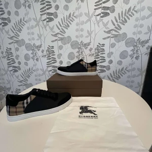 Burberry sneakers  - Snygga sneakers från Burberry med klassiskt beige, svart och röd rutigt mönster på hälen och sidorna. Skorna har svart ovandel, vita platta sulor och svarta snören. Perfekta för dig som gillar stilrena och exklusiva sneakers. OBS! PRISET KAN DISKUTERAS VID EN SNABB AFFÄR!