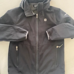 Svart skaljacka från 8848 altitude - Svart skaljacka med huva från 8848 altitude. Jackan har dragkedja framtill och två sidofickor med dragkedjor.tryck på bröstet och logga på ärmen. Perfekt för kyliga dagar.Skal jacka 8848 Altitude Litet hål i yttertyget, syns mkt lite på jackan storlek 160