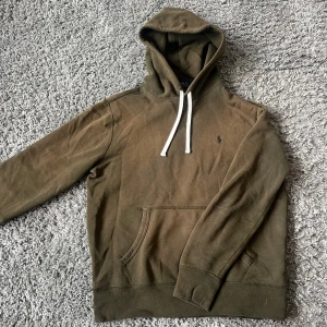 Olivgrön hoodie från Polo Ralph Lauren - Säljer en olivgrön hoodie från Polo Ralph Lauren med vit dragsko och klassisk känguruficka. Tröjan är i bra skick ser lite missfärgad ut men den är ej de i verkligeheten.
