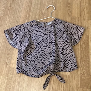 Prickig blus med knyt från Lindex Kids - Säljer en luftig och söt blus från Lindex Kids i storlek 128. Blusen är lila med svarta prickar, har korta vida ärmar och knyt framtill. Perfekt för varma dagar och enkel att matcha med jeans eller shorts. Några få fläckar så säljer billigt. 