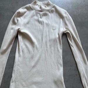 Beige ribbad polotröja från Lee - Säljer en beige ribbad polotröja från Lee med diskret broderad logga på bröstet. Tröjan har lång ärm och en hög krage,  Mjuk och stretchig i materialet.