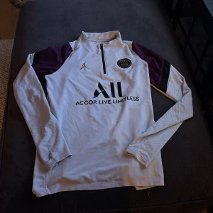 Vit långärmad PSG fotbollströja från Jordan - Säljer en vit långärmad PSG fotbollströja från Jordan med svarta och vinröda detaljer på axlarna. Tröjan har halv dragkedja, PSG-märke på bröstet och Jordan-logga i guld. Materialet är lätt och andas.