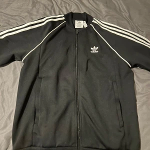 Svart Adidas track jacket med vita ränder - Klassisk svart Adidas track jacket med vita ränder längs ärmarna och dragkedja framtill. Jackan har Adidas-logga på bröstet och två fickor. Perfekt för en sportig och avslappnad stil.