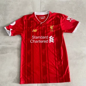 Liverpool FC Salah 11 matchtröja - Röd Liverpool FC matchtröja med vita detaljer, tryck av M. Salah och nummer 11 på ryggen. Tröjan har klubbmärke, sponsorlogga och New Balance-logga på bröstet. Klassisk V-ringning och korta ärmar. Materialet är lätt och andas, perfekt för fotbollsträning eller match.