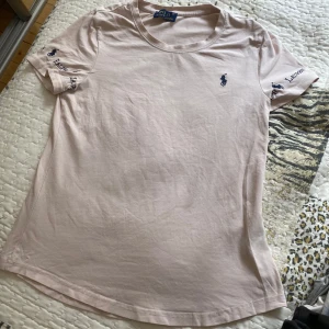 Ljusrosa t-shirt från Ralph Lauren - Snygg ljusrosa t-shirt från Ralph Lauren med klassisk blå broderad logga på bröstet och broderade detaljer på ärmarna. T-shirten har rund halsringning och är tillverkad i mjuk bomull. Perfekt för en clean och stilren look.