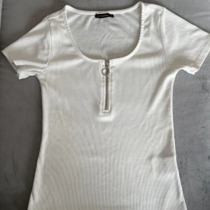 Vit ribbad topp med dragkedja - Vit ribbad topp från med korta ärmar och rundad halsringning. Toppen har en snygg silverfärgad dragkedja framtill med ringdetalj. Figurnära passform och stretchigt material som känns bekvämt och trendigt.