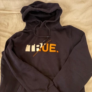 Svart hoodie med TRUE-tryck - Svart hoodie med huva och snörning. STORLEK M. På bröstet finns ett stort tryck och brodyr i vitt och orange med texten TRUE och en diagonal linje. Klassisk känguruficka framtill och långa ärmar. Perfekt för en chill och avslappnad stil. Hör av er vid frågor och intresse!