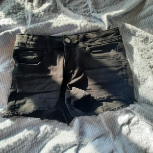 Svarta jeansshorts med fransig kant - Svarta jeansshorts med rå, fransig kant och klassisk femficksmodell. De har låg midja och är perfekta för varma dagar. Snygg, enkel design som passar till det mesta. Dragkedja och knapp framtill.
