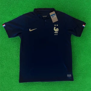Frankrike Nike fotbollströja marinblå - Snygg marinblå Frankrike fotbollströja från Nike med guldiga detaljer, broderad tupp och två stjärnor på bröstet. Kortärmad modell med liten krage och fransk flagga på ärmarna. Tillverkad i lätt och ventilerande material, perfekt för match eller träning.