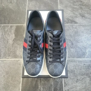 Gucci sneakers med röd och blå rand - Säljer ett par snygga Gucci sneakers i mörkblått med GG-mönster och klassiska röda och blå ränder på sidorna. Skorna har svarta snören, vit sula och rund tå. Perfekta för dig som gillar exklusiva märken och stilren design.