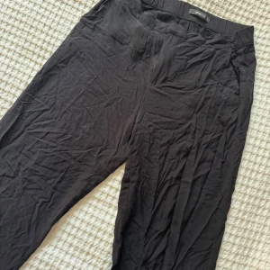 Svarta byxor från Gina Tricot - Svarta byxor från Gina Tricot med normal passform och hög midja. Byxorna har dragkedja och knapp framtill samt fickor på sidorna. Perfekta för en stilren och enkel look.