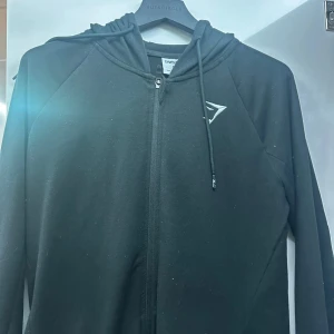 Svart hoodie från Gymshark - Säljer en svart hoodie från Gymshark i storlek M. Den har en dragkedja framtill och en liten logga på bröstet. Perfekt för träning eller en avslappnad dag. Huvan har snören för justering. Skön och stilren design!