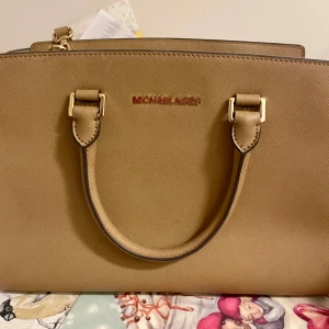 Beige handväska från Mich@el Kors - Snygg beige handväska från Mich@el Kors med guldfärgade detaljer och dubbla handtag. Väskan har dragkedja upptill och flera fack inuti för smart förvaring. Perfekt accessoar för att lyfta din outfit.