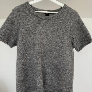 Grå mohair stickad kortärmad tröja  - Mysig grå stickad tröja i mohair (kvalitetsull, mycket mjuk) från Lindex med korta ärmar och rund halsringning. Perfekt för lager-på-lager eller som en enkel bas i garderoben. Mjuk och bekväm, passar till många olika stilar. Nopprar inte. Aldrig använd.