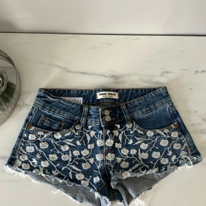 Blommiga jeansshorts från tally weijl  - Snygga blå jeansshorts med vit blommig brodyr framtill och råa fransade benslut. Klassisk femficksmodell med dubbla knappar i midjan. Perfekta för sommaren! Nya utan prislapp