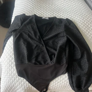 Svart glittrig omlott bodysuit från NA-KD - Säljer en svart glittrig bodysuit från NA-KD med omlott-design och v-ringning. Långa ärmar och åtsittande passform, perfekt för att sticka ut. Superfin till fest eller när du vill glänsa lite extra ✨