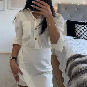 Vit skjortklänning från Zara - Vit skjortklänning från Zara med långa ärmar, krage och dekorativa träknappar framtill. Klänningen har en markerad midja och en bröstficka, samt en asymmetrisk detalj vid kjolen.  Helt ny med prislappen kvar