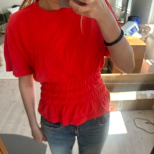 Röd topp med smockad midja - Säljer en röd topp med korta ärmar och smockad midja som ger en snygg form. Toppen har en rund halsringning och är tillverkad i ett mjukt material. Perfekt för att lyfta vilken outfit som helst.