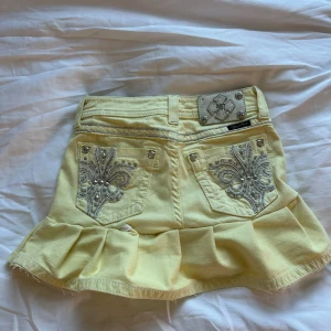 Gul volangkjol från Miss Me - Supersöt gul kortkjol från Miss Me med volangkant och snygga broderade detaljer med pärlor på bakfickorna. Kjolen har jeansmaterial och dekorativa nitar. Perfekt för dig som vill sticka ut lite extra!