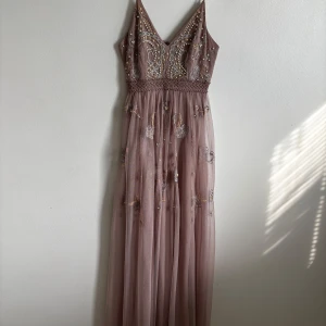 Lång lila klänning med pärlor och broderi - Drömmig långklänning i lila mesh med tunna axelband, v-ringning och vackra broderade blommor. Överdelen är dekorerad med pärlor och silvriga detaljer, och kjolen har broderade blommor med pärlor. Perfekt för bal eller festligheter.