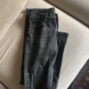 Svarta jeans från Nudie Jeans - Säljer ett par svarta jeans från Nudie Jeans med klassisk femficksdesign och snygga detaljer på bakfickorna. Jeansen har en något tvättad look och raka ben. Perfekta för dig som gillar stilrena och bekväma jeans.