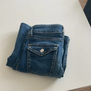 Blå bootcut jeans med fickdetaljer - Snygga blå jeans med bootcut passform och coola bakfickor med lock och knapp. Klassisk denim med kontrastsömmar och normal midja. Perfekta för dig som gillar en tidlös jeanslook med lite extra detaljer.