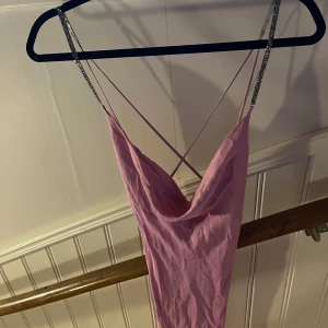 Rosa kortklänning med strassband - Supersöt rosa kortklänning med tunna axelband och glittriga strassdetaljer på banden. Klänningen har en snygg draperad urringning och öppen rygg med korsade band. Perfekt för dig som vill sticka ut och glänsa lite extra.