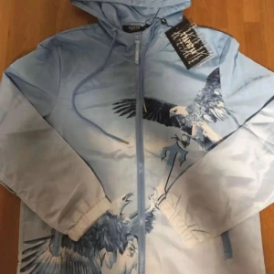 Trapstar blå vindjacka med örntryck - Snygg blå vindjacka från Trapstar med huva och dragkedja. Jackan har ett fett örntryck över bröstet och ärmarna, samt snörning vid huvan. Perfekt för dig som vill sticka ut med streetwear-vibe.