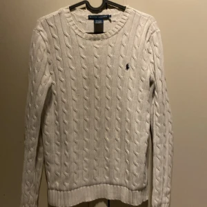 Vit kabelstickad tröja från Ralph Lauren - Vit kabelstickad tröja från Ralph Lauren med klassisk rund halsringning och broderad marinblå logga på bröstet. Tröjan är långärmad och har ribbade muddar vid ärmslut och nederkant. Perfekt för en clean och stilren look. 