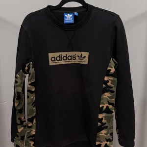 Svart Adidas tröja med camouflageärmar - Svart långärmad tröja från Adidas med stort logotryck framtill och ärmar i grön camouflage. Tröjan har klassisk rund hals och är gjord i mjuk bomullsmix. Perfekt för dig som gillar streetwear och vill sticka ut lite extra.