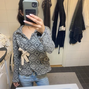 Odd Molly - Säljer en grå och vit cardigan jacka med rutigt mönster och dekorativ rosett i beige på sidan. Jackan har v-ringad hals och långa ärmar, perfekt för dig som gillar unika detaljer och mysig stil.