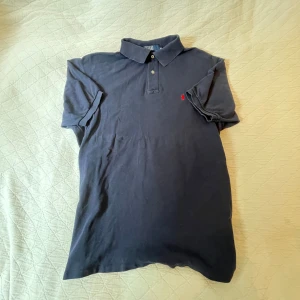 Mörkblå piké Polo Ralph Lauren - Klassisk mörkblå pikétröja från Polo Ralph Lauren i 100% bomull. Tröjan har korta ärmar, krage med två knappar och den ikoniska röda logotypen broderad på bröstet. Perfekt för en clean och tidlös stil.