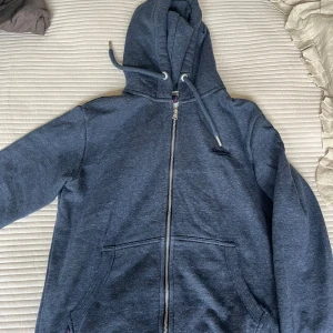 Mörkblå hoodie från Superdry M - Mysig mörkblå hoodie från Superdry med dragkedja och huva. Tröjan har stora fickor framtill, snörning i huvan och är fodrad med mjuk fleece på insidan. Perfekt för kyliga dagar och chill vibes.