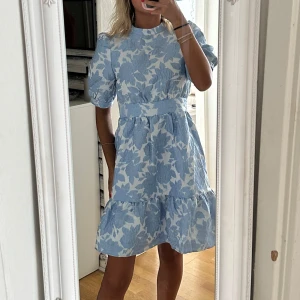 Blå blommig klänning med volang - Nyköpt men går inte att retunera, beställde fel storlek. Supersöt blå klänning med stora blommor och volang nertill. Klänningen har korta puffärmar och markerad midja. 