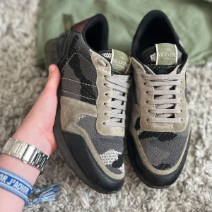 Valentino Rockrunner sneakers i camouflage - Säljer ett par Valentino Rockrunner sneakers med grått och svart camouflage-mönster, beige mockadetaljer och svarta partier. Skorna har snörning, rund tå och en robust gummisula med nitar på hälen. Perfekta för dig som gillar streetstyle och exklusiva märken.