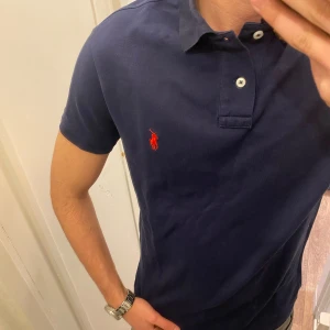 Mörkblå Piké från Polo Ralph Lauren - Säljer en klassisk mörkblå pikétröja från Ralph Lauren med röd broderad logga på bröstet. Tröjan har korta ärmar, krage och knappar vid halsen. Perfekt för en stilren och avslappnad look. Storlek Medium Classic Fit. Snabb Leverans!