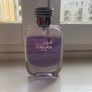 Lila parfym Hawas For Him - En stilren parfymflaska med lila vätska och silverfärgad kork. Flaskan har raka linjer och tydlig etikett med texten 'Hawaf For Him' på framsidan. Perfekt för dig som gillar eleganta och moderna dofter.