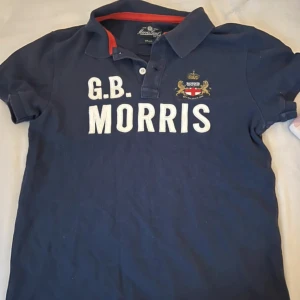 Marinblå pikétröja från Morris - Säljer en marinblå pikétröja från Morris med vit broderad text 'G.B. MORRIS' och ett broderat emblem på bröstet. Tröjan har klassisk krage, knappar framtill och är tillverkad i 100% bomull. Perfekt för en stilren och sportig look.